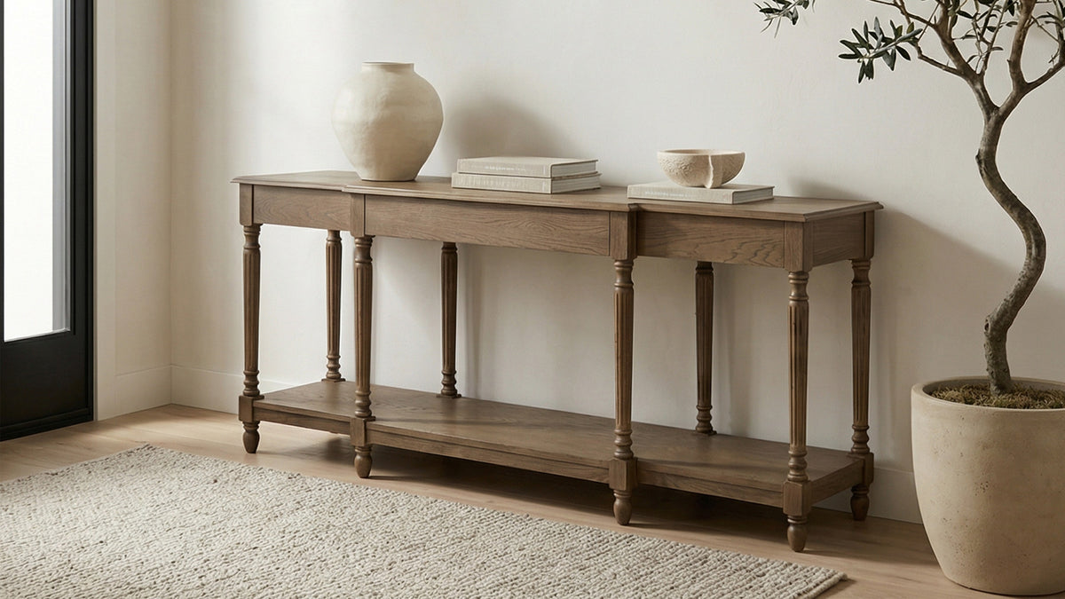 Moe's Home Collection Martha console table in limed oak, its turned legs and lower shelf seen against a white wall, styled with a rounded vase, stacked books and a small bowl beside a potted olive tree