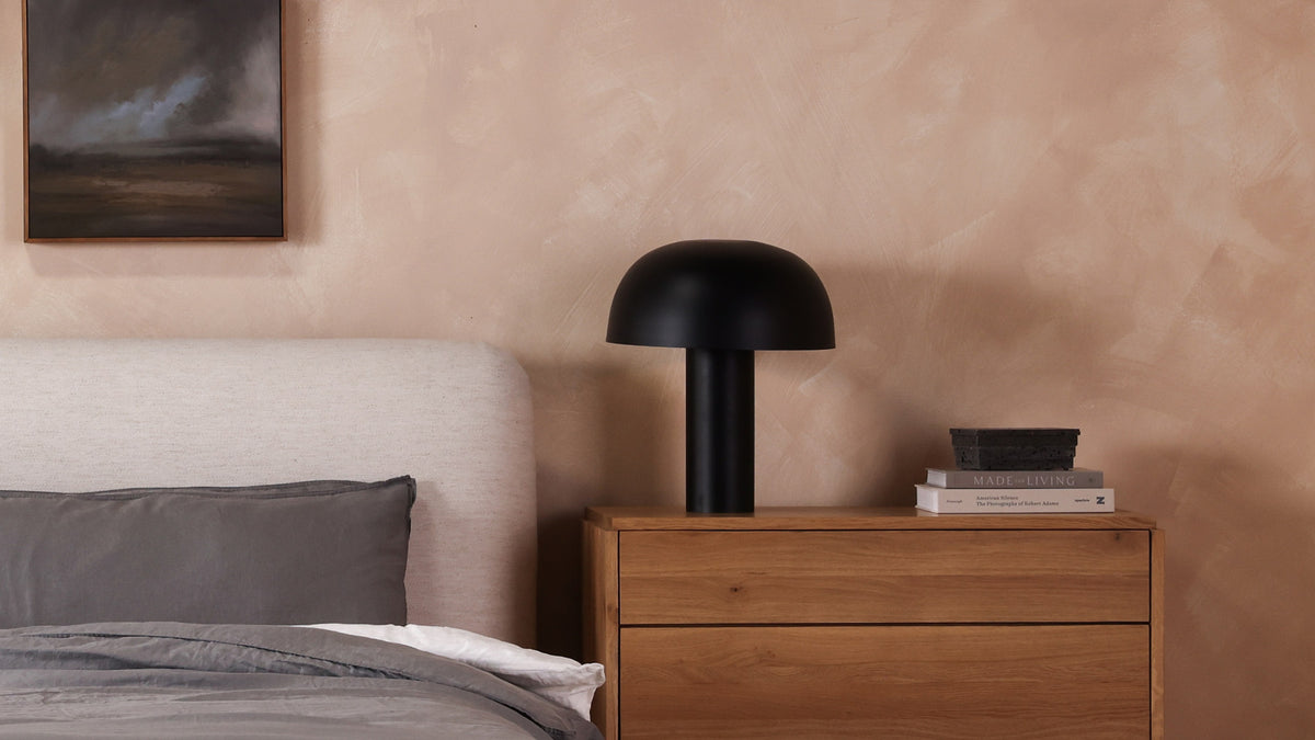 The Nanu table lamp in black on an oak nightstand beside an upholstered bed, its domed iron shade over a slim column, with a stone box on stacked books and a dark landscape painting above