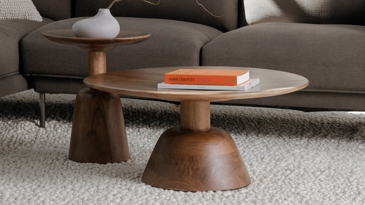Moe's Home Collection Nels coffee table in dark brown acacia, its round top and bell-shaped pedestal base on an ivory looped wool rug before a deep brown sectional, with the matching Nels end table alongside