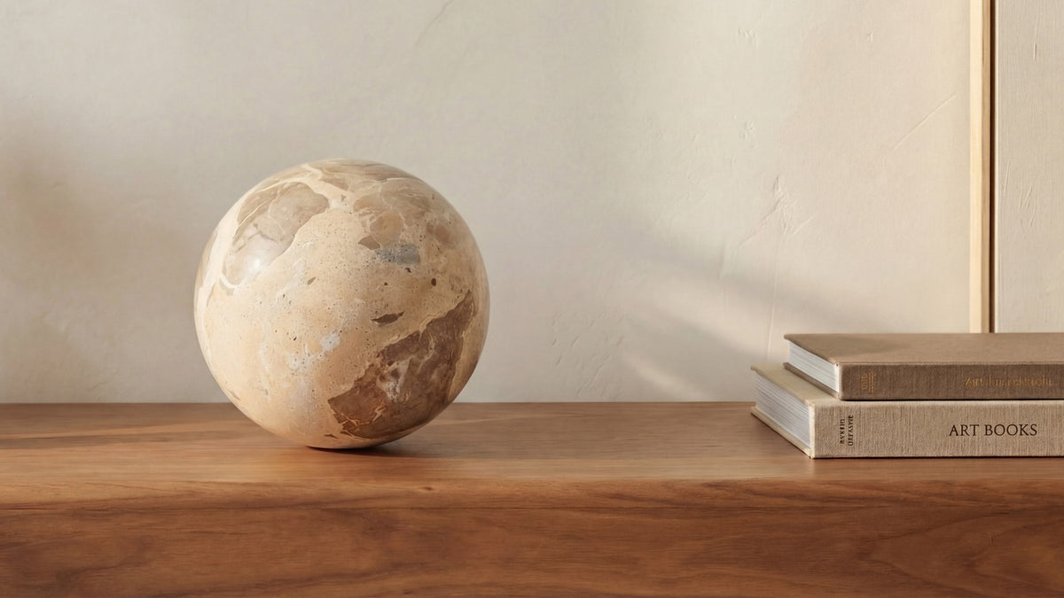 The Odessa sphere in tendu beige marble on a walnut surface beside a stack of art books, its mottled brown and cream veining in raking light