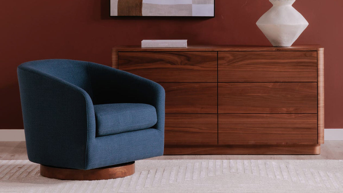 A Moe's Home Collection Oscy swivel chair in deep navy Dark Atlantic upholstery on a dark wooden base, placed at the left of a room with an oxblood wall, a walnut sideboard, an abstract painting and a white vase of dried grasses