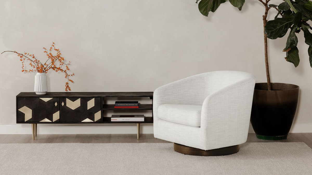 A Moe's Home Collection Oscy swivel chair in flecked cream Splashed White upholstery beside a black and bone patterned media console and a fiddle-leaf fig, against a pale plaster wall