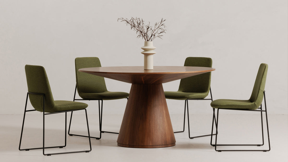 Moe's Home Collection Otago round dining table in walnut on a pale studio floor, the cone-shaped pedestal base rising to a flared underside beneath the round top, with four moss green upholstered chairs on black sled frames and a pale ceramic vase of dried branches at the center