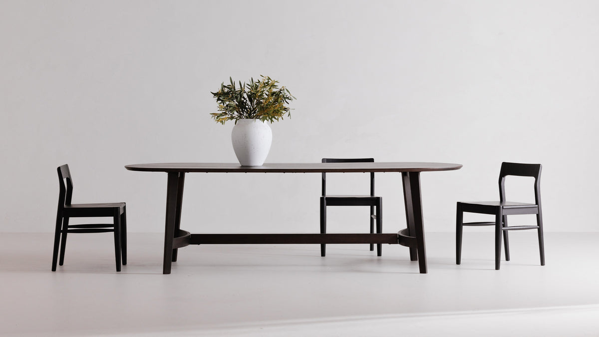 Three Moe's Home Collection Owing dining chairs in black solid oak around a dark oval dining table in a white studio, the open backs framing the table legs behind