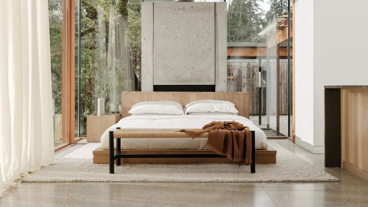 Moe's Home Collection Plank bed in natural oak with a wide plank headboard, made up in white bedding on a cream rug in a glass-walled room with concrete walls and forest beyond