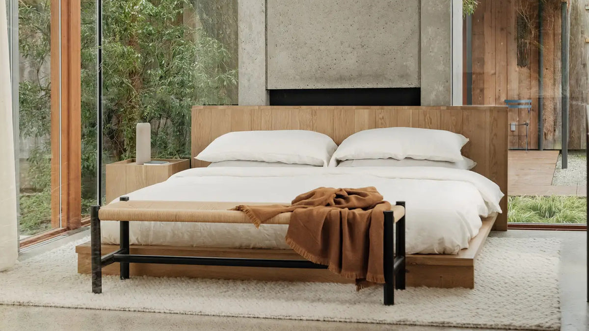 The Plank bed seen closer, with a rust throw over a black bench at the foot and the oak headboard running the full width behind white bedding