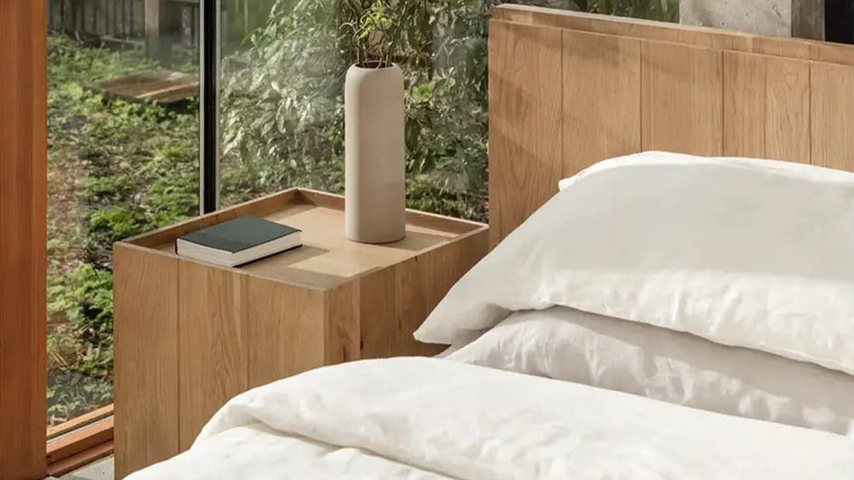 Close view of the Moe's Home Collection Plank nightstand in natural oak, a book and a tall cream vase resting inside the raised rim of its lipped tray top, with the garden through the glass behind