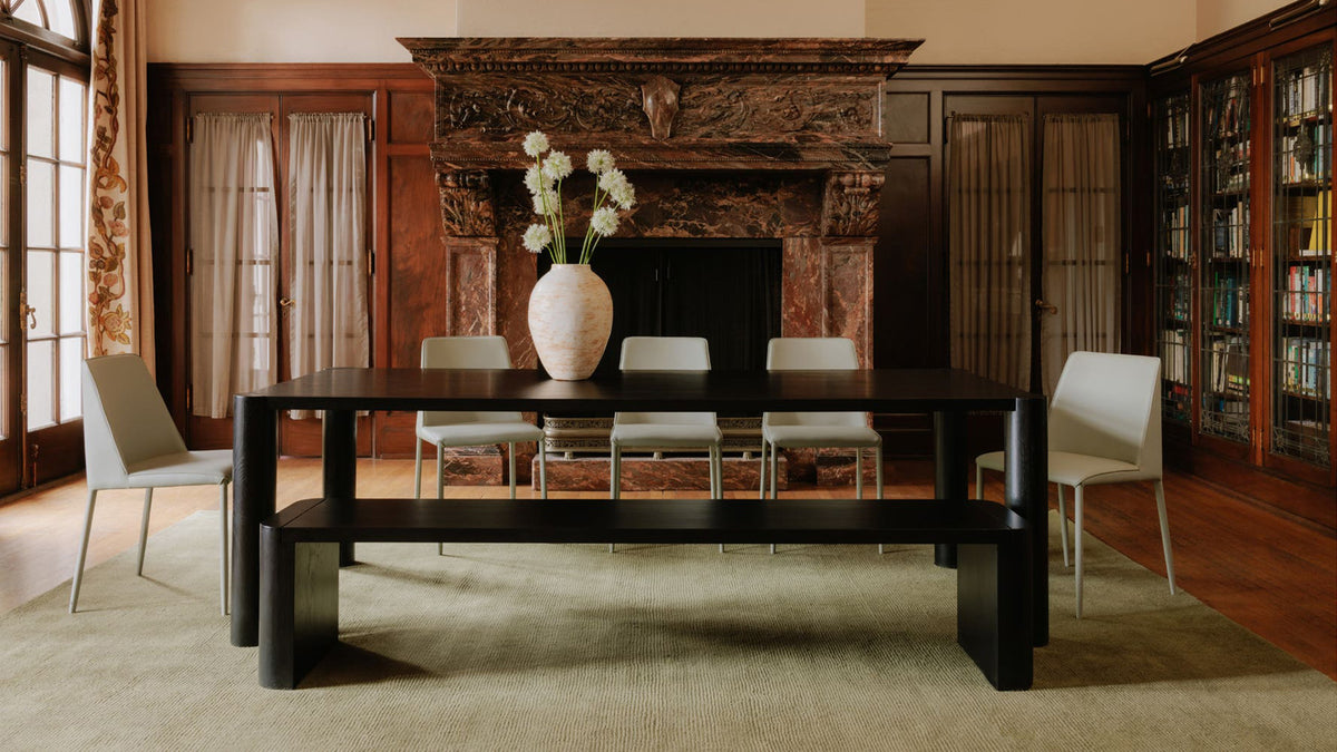 Two Moe's Home Collection Post dining benches in blackened solid oak drawn up to a dark dining table in a panelled room with a carved stone fireplace