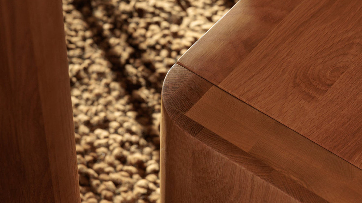 Close view of the Post dining bench in natural solid oak, showing the rounded corner where the slab seat turns down into the leg