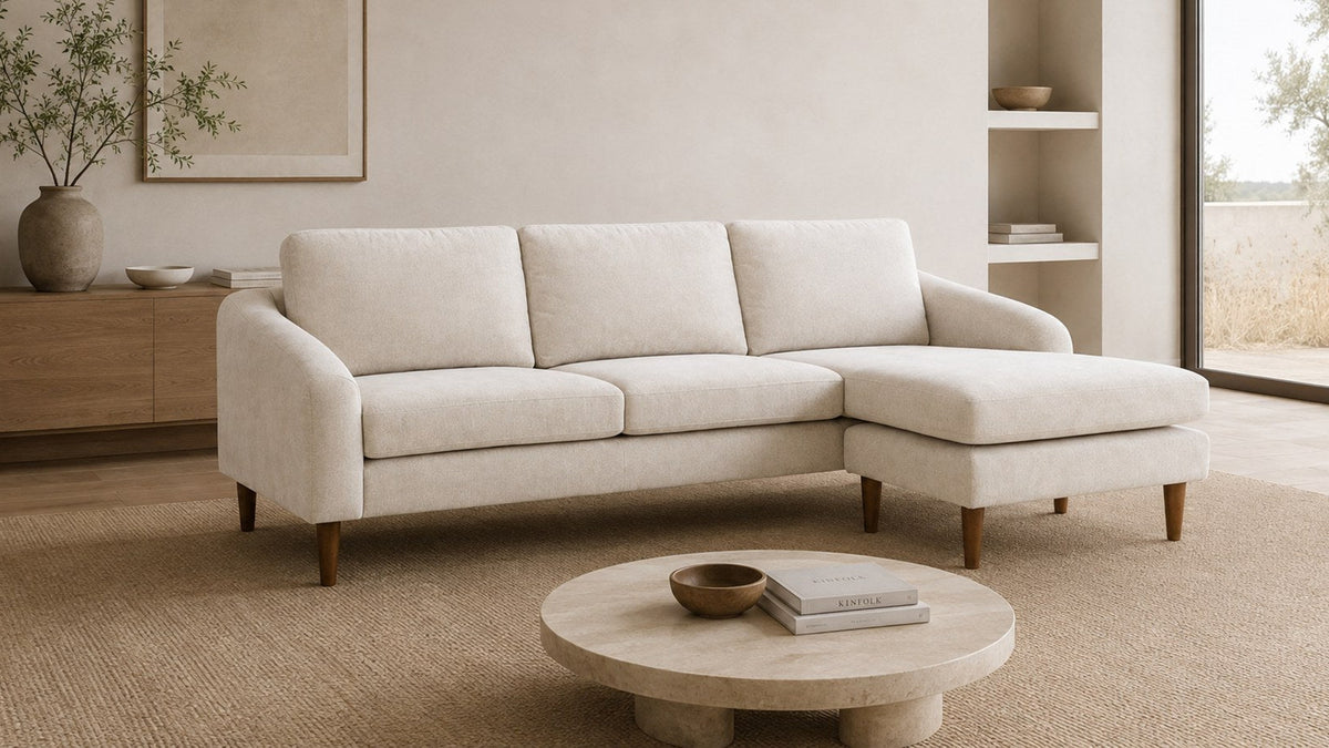 Moe's Home Collection Quinn Sectional in oatmeal with the chaise ottoman on the right, seen at an angle on a jute rug with a round stone coffee table in front, an oak sideboard and framed art to the left and a glass wall onto dry grass at the right