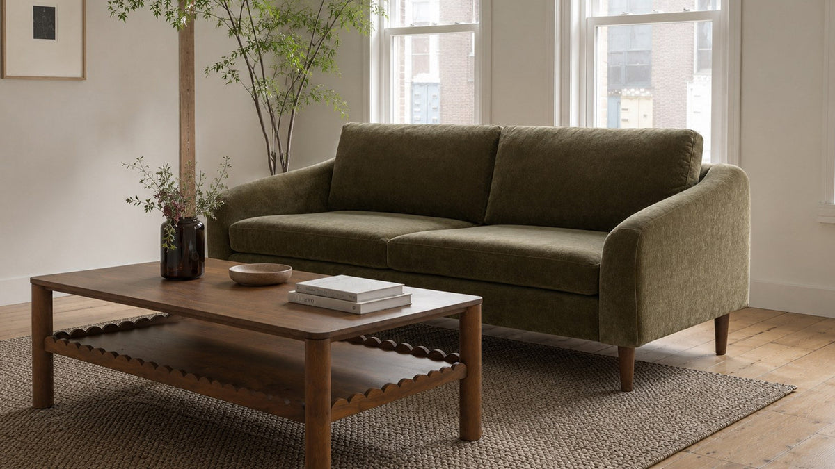 Moe's Home Collection Quinn sofa in cedar green, its low sloped arms and tapered wood legs seen at an angle in a bright room with tall sash windows, a small maple in a planter behind it and a walnut coffee table on a jute rug in front