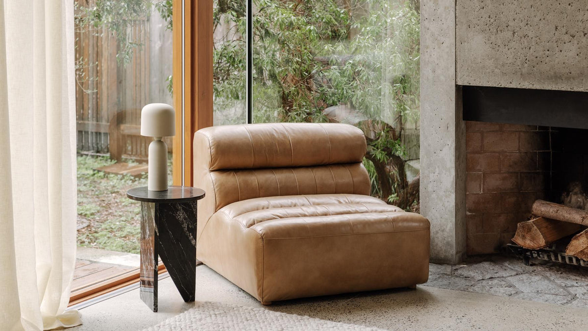 Moe's Home Collection Ramsay slipper chair in tan top-grain leather, armless with a low channeled back, standing on a polished concrete floor beside a sliding glass door and a board-formed concrete fireplace, with a black marble side table and a small lamp to its left