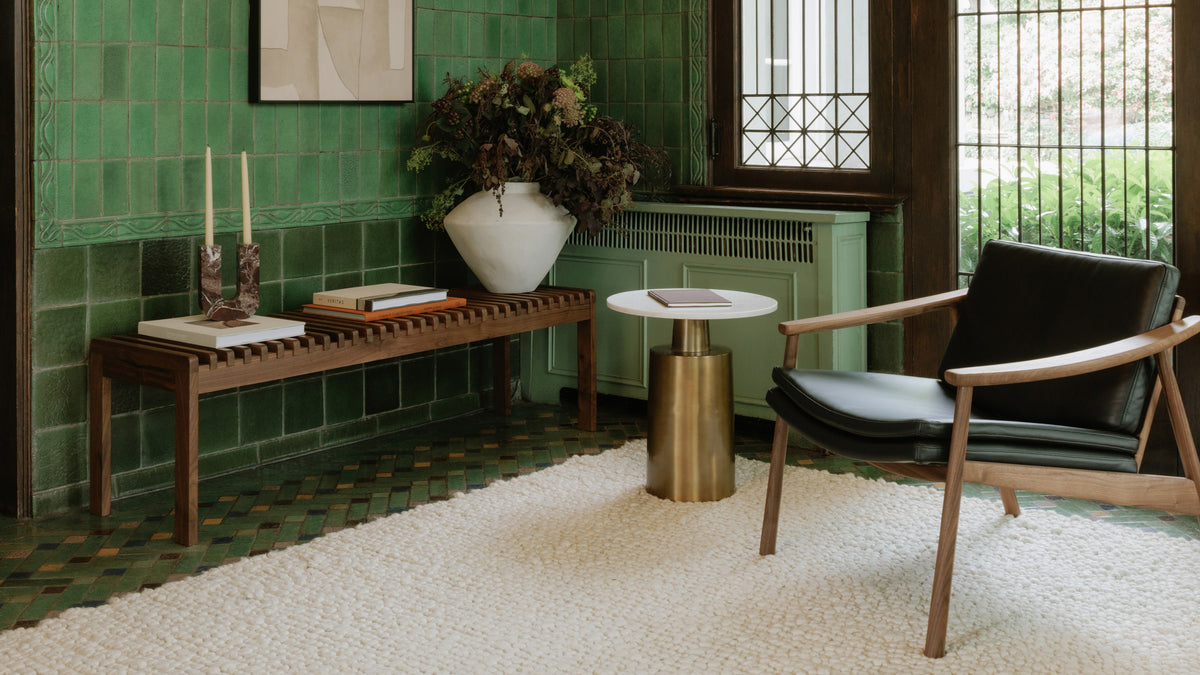 Moe's Home Collection Rohe bench in solid walnut, its slatted seat stacked with books, in a green-tiled nook beside a low chair and a round side table