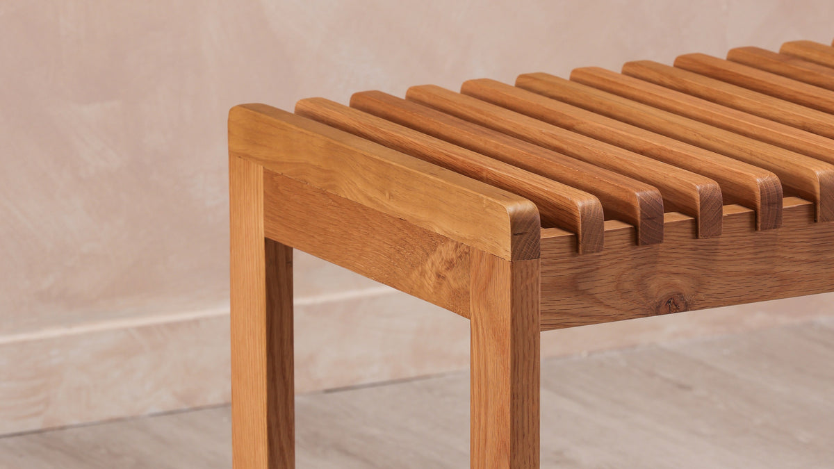 Close view of the Rohe bench in solid oak, showing the run of spaced slats across the seat and the square leg at the corner