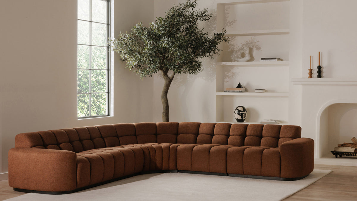 Moe's Home Collection Roman L-shaped sectional in rust, its square-tufted seat and back running the full width of a pale plaster room, with an olive tree and built-in shelving behind it and a fireplace to the right
