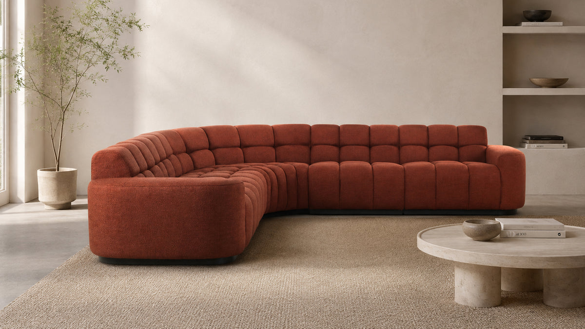 Moe's Home Collection Roman L-shaped sectional in rust on a woven rug, seen straight on with its curved corner and both runs visible, beside a round travertine coffee table and a potted tree