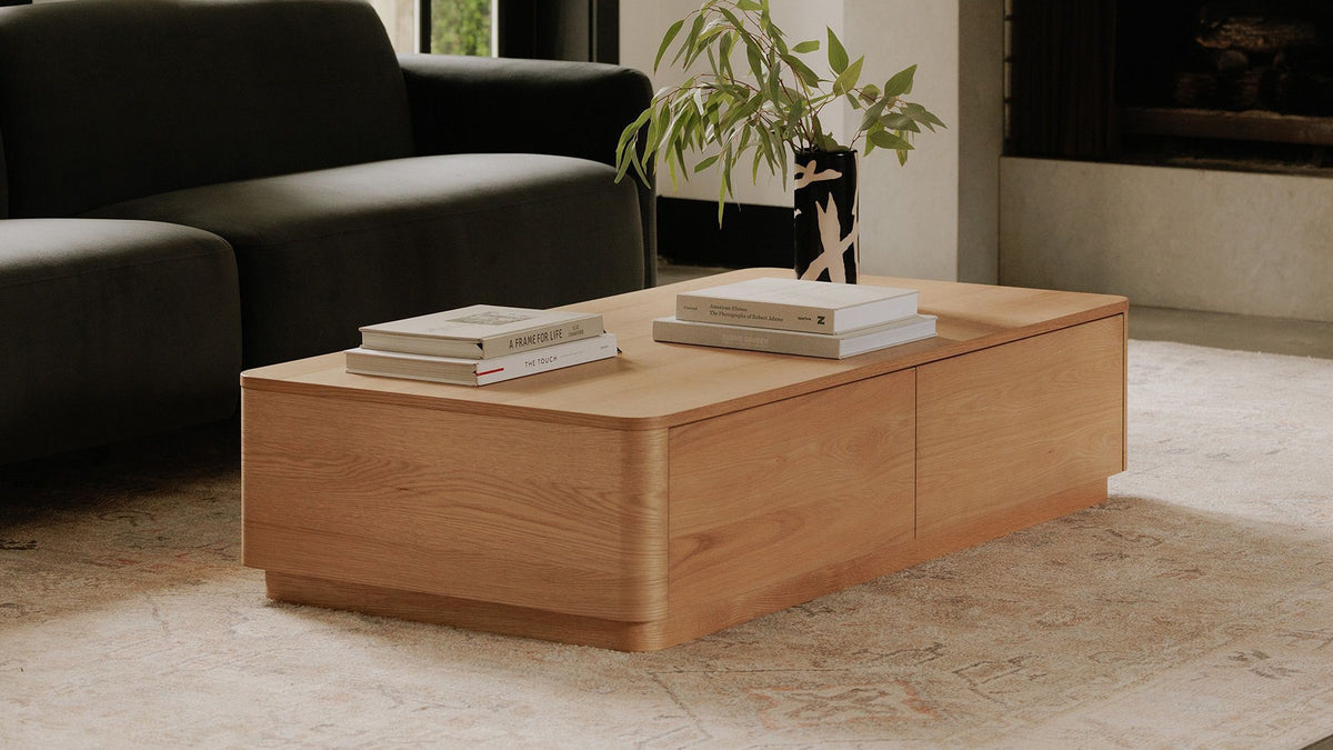 Moe's Home Collection Round Off coffee table in natural oak, styled with stacked art books and a branch in a black vase on a pale rug beside a dark green sofa