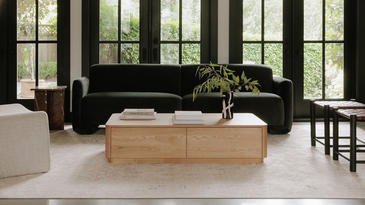 Moe's Home Collection Round Off coffee table seen head on in a living room with black steel doors, a dark green sofa and a woven leather bench