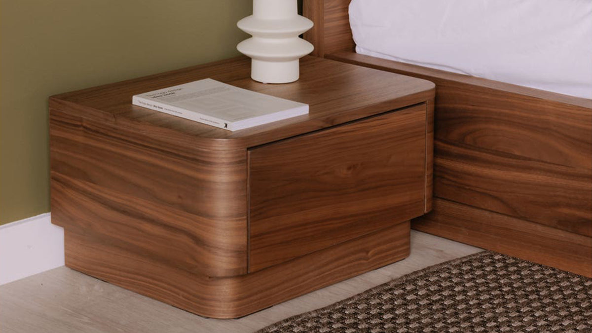Moe's Home Collection Round Off Small nightstand in walnut brown, a single deep drawer in a rounded case on a recessed plinth, standing low beside a walnut bed frame against an olive wall