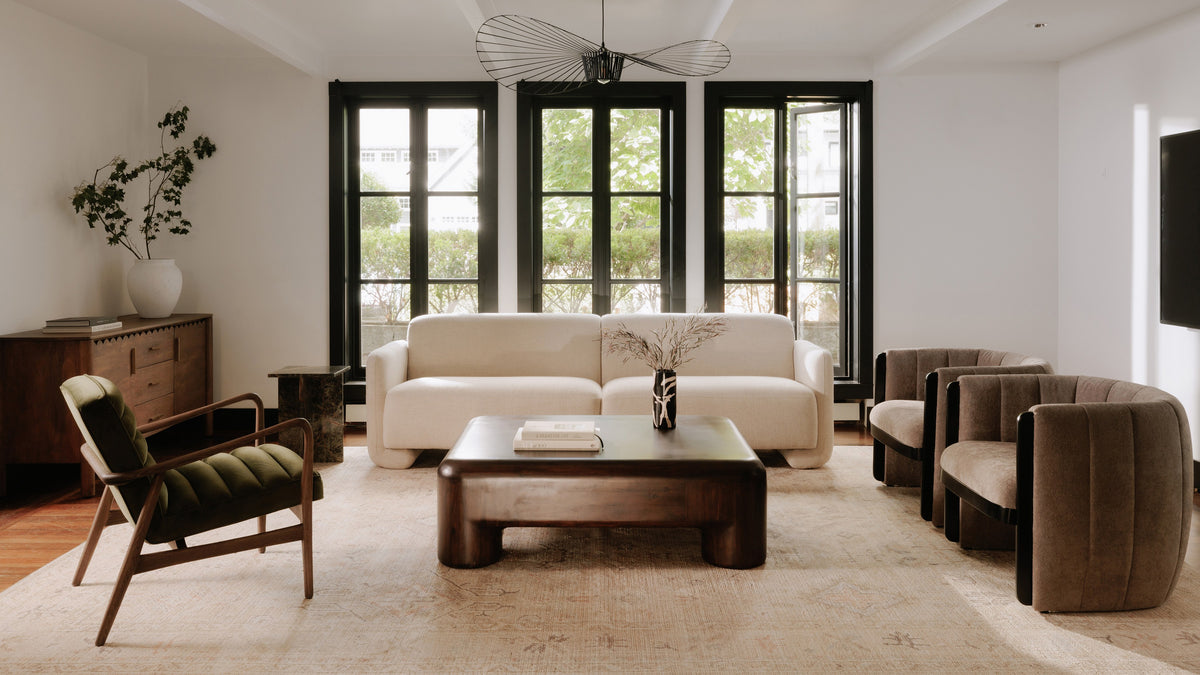 Moe's Home Collection Rowan coffee table in dark brown stained acacia, centred on a faded patterned rug in a bright living room with a cream boucle sofa, two taupe channel-tufted swivel chairs, a green velvet lounge chair and tall black-framed windows
