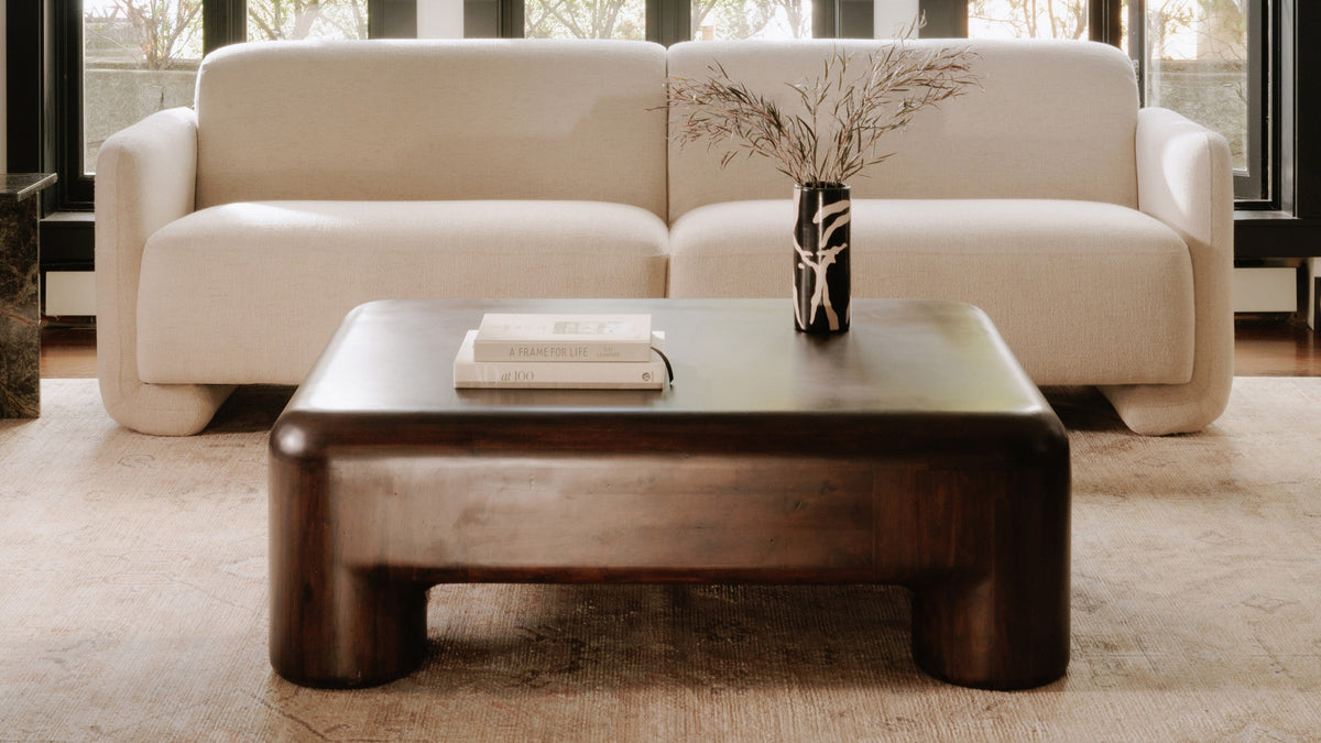 Closer view of the Rowan coffee table's rounded dark acacia form in front of a cream boucle sofa, with two art books and a black and white patterned vase of dried branches on the top, and the open arch between its cylindrical corner legs visible beneath