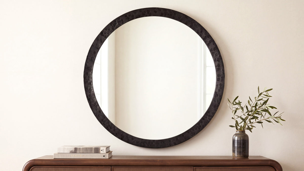 The large Royce mirror in black above a walnut console, its hammered iron rim reading as texture against a plain wall, beside a small vase of olive