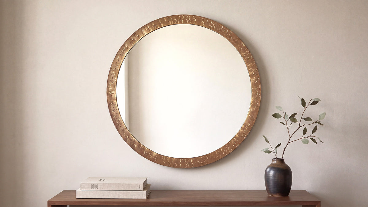 The small Royce mirror in bronze above a wooden console, a stack of books to one side and a dark vase with a single leafed stem