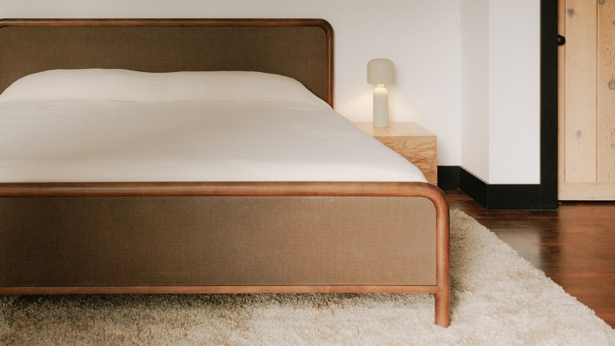The Rye bed seen from the foot, showing the rounded corner posts and the woven footboard panel against white bedding and a dark wood floor