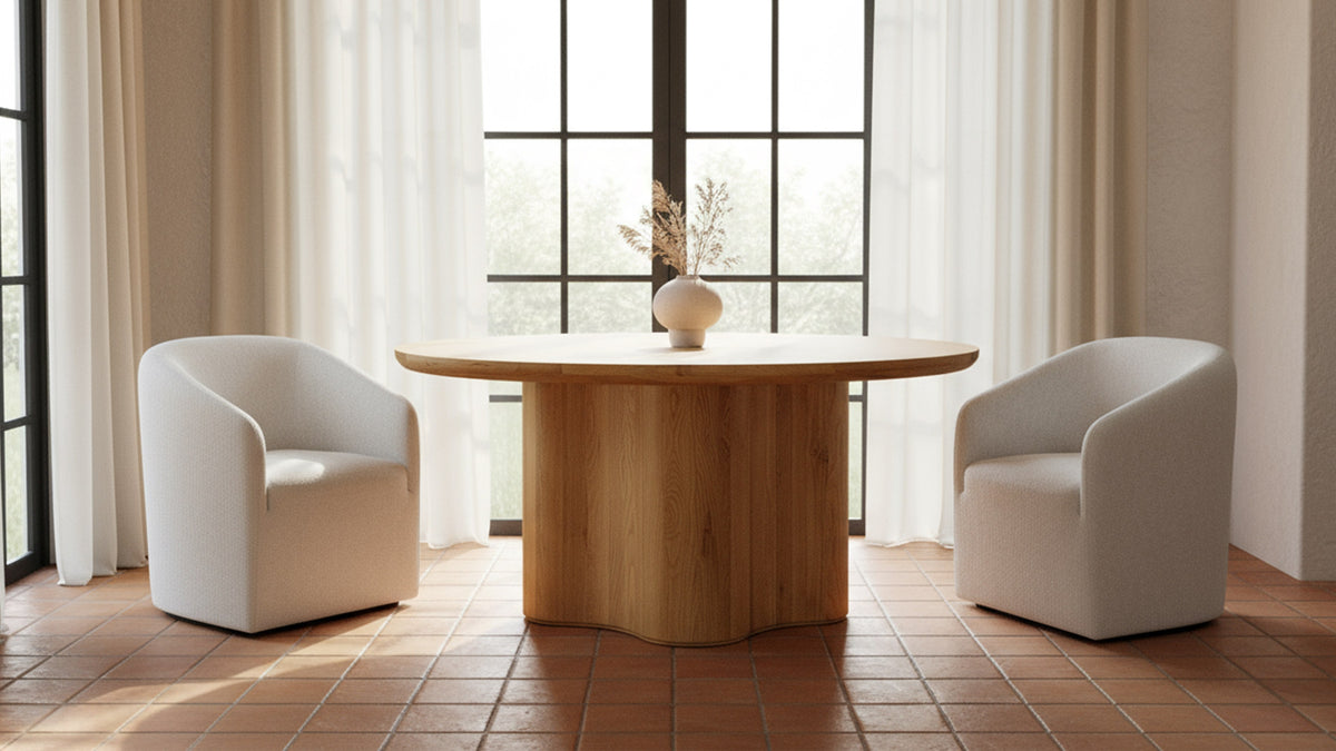 Moe's Home Collection Sidney dining table in Natural oak, its round top carried on a single continuous oak base, standing on a terracotta tile floor between two ivory upholstered armchairs, with a tall black-framed window and sheer curtains behind it