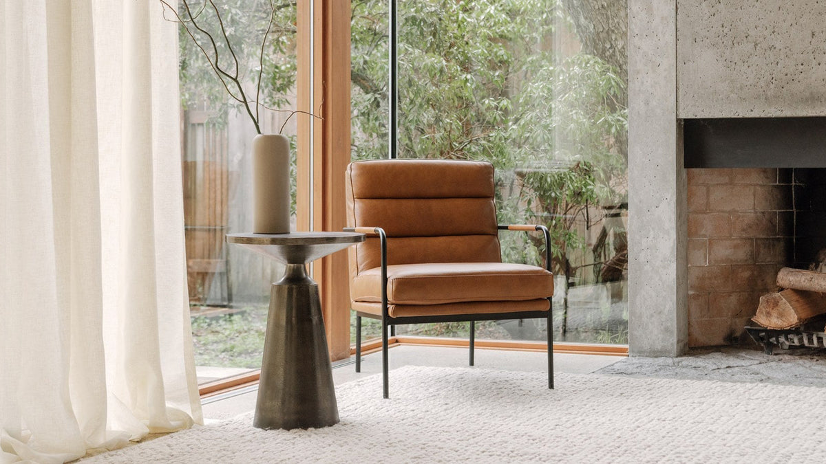 Moe's Home Collection Sonja accent table in charcoal aluminum, its cast pedestal base beside a tan leather lounge chair on a wool rug, with a tall ceramic vase on the round top