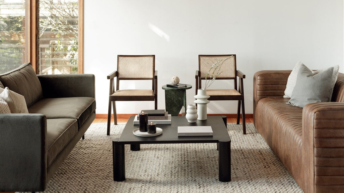 A pair of Moe's Home Collection Takashi chairs in dark brown elm with woven rattan seats and backs, set side by side in a living room between an olive sofa and a brown leather sofa, with a low black coffee table on a pale rug