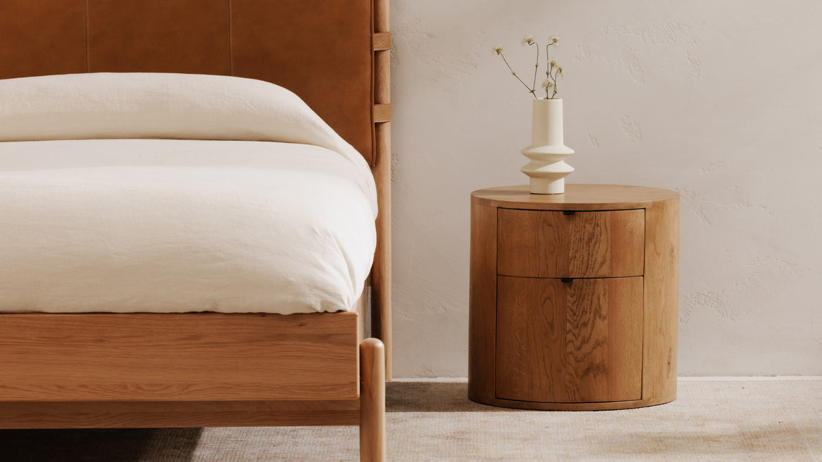 Moe's Home Collection Theo 2-Drawer nightstand in natural oak beside a bed with a tan leather headboard, its round drum case closed to the floor with two curved drawer fronts and a white sculptural vase on the solid top