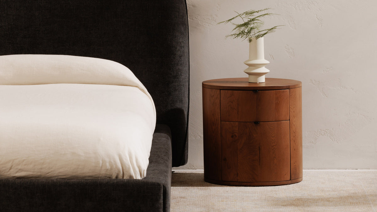 Moe's Home Collection Theo 2-Drawer nightstand in brown oak against a plaster wall beside a charcoal bed, the round case closed all the way to the floor with two curved drawer fronts and a white sculptural vase above