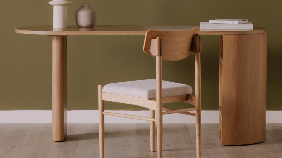 Moe's Home Collection Theo solid oak desk against an olive wall, its oval top and rounded pedestal holding a lamp, a vase and a stack of books, with a wooden chair pulled up to it