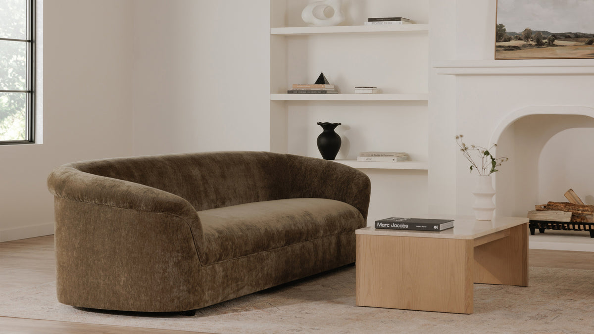Moe's Home Collection Thora sofa in Dark Green, its continuous rolled edge and floor-meeting base seen against a pale plaster wall, beside a low light-oak and stone coffee table on a soft rug