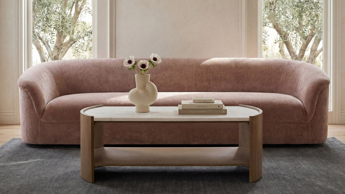 Moe's Home Collection Thora sofa in Blush, shown straight on between tall windows with olive trees beyond, behind a marble-topped oak coffee table on a gray rug
