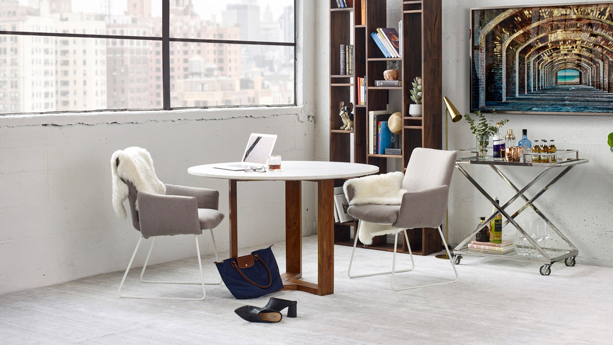 Moe's Home Collection Trumpet floor lamp in brass, its slim stem arching over a mirrored bar cart in a loft dining corner with a round white table, grey sled-base chairs, a tall walnut bookcase and city windows