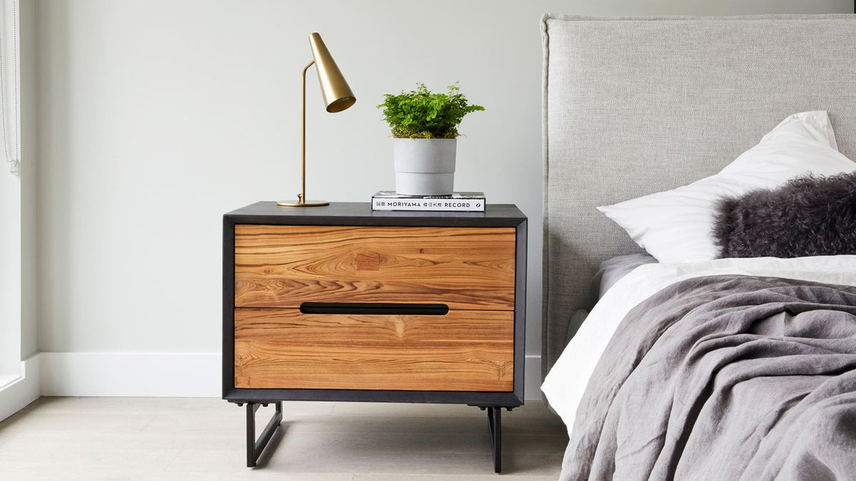 The Trumpet table lamp in brass on a walnut and black nightstand beside a grey upholstered bed, its narrow cone shade angled over a book and a small potted plant