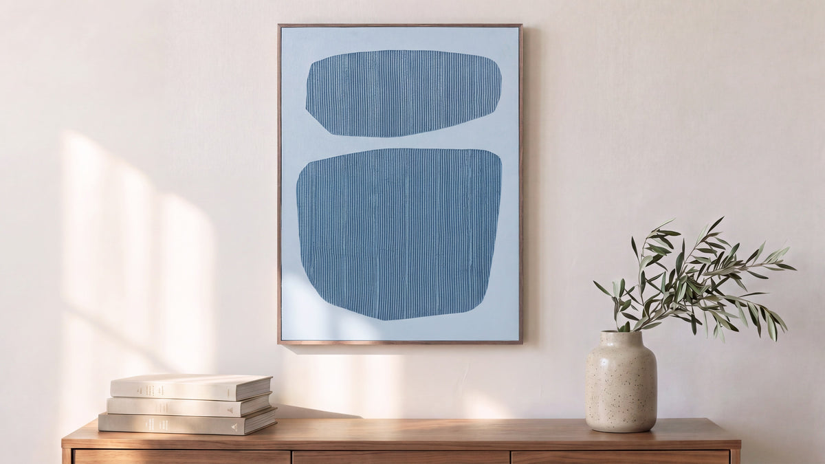 The Uma framed painting leaning against a sunlit wall above a walnut sideboard, its two blue forms on pale ground, beside a stone vase of olive branches and stacked books