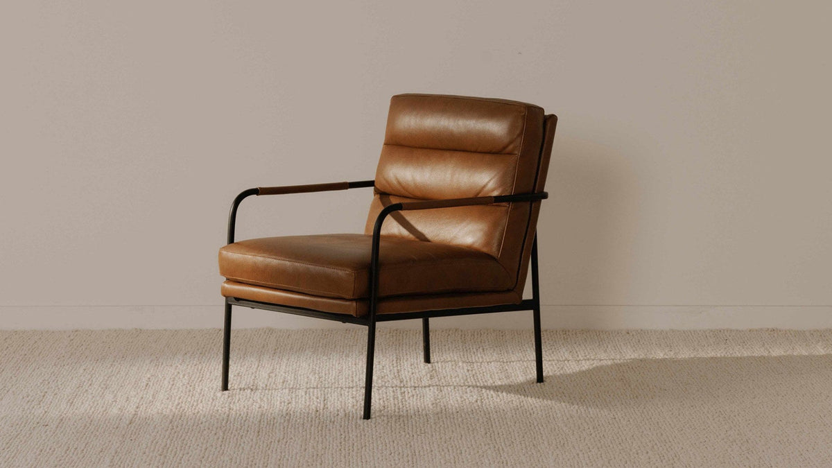 A Moe's Home Collection Verlaine armchair in chestnut brown top-grain leather, three-quarter view on a textured cream rug against a plain warm gray wall, showing the channelled back cushion and the leather-wrapped arm rail