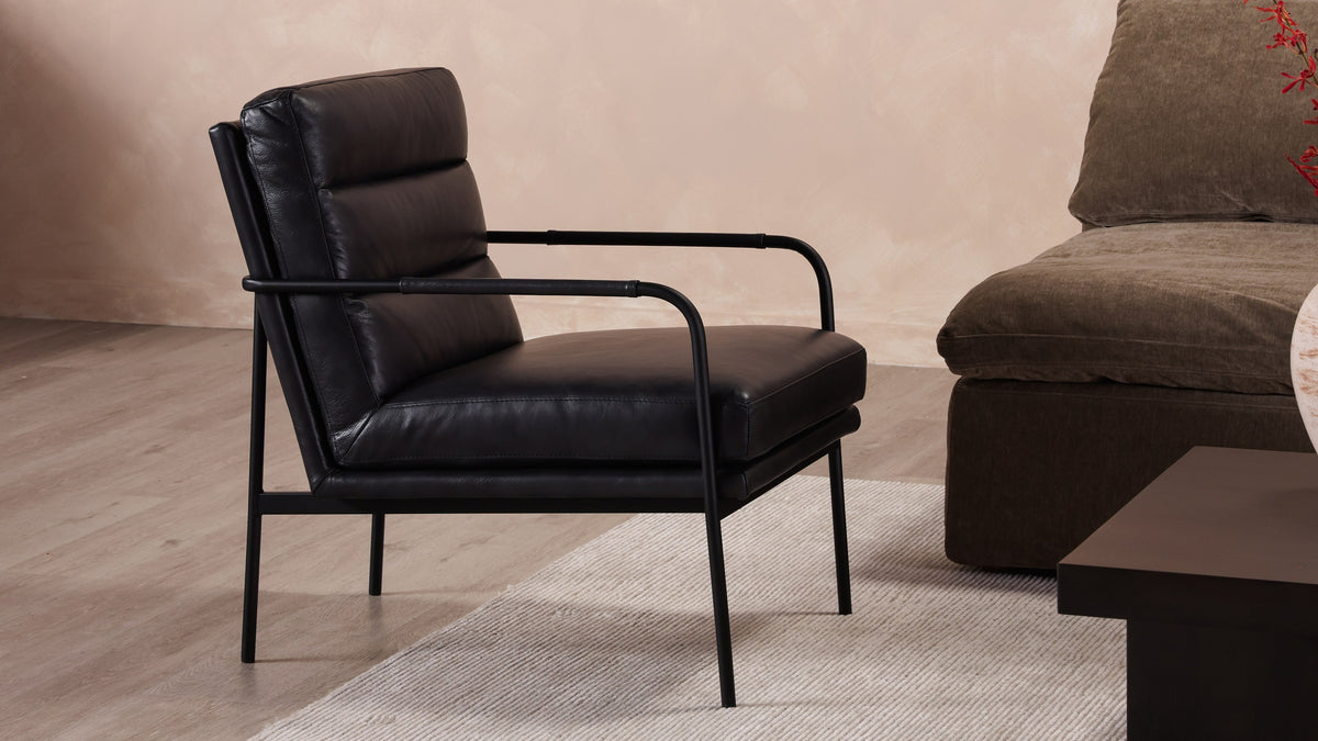 A pair of Moe's Home Collection Verlaine armchairs in raven black top-grain leather at the left of a living room, angled toward an olive-brown sectional with a dark wood coffee table between them and three framed artworks on a plaster wall