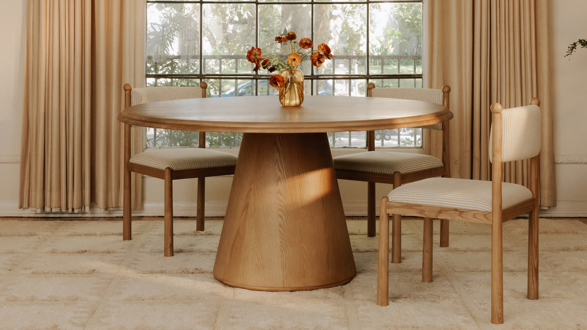 Three Moe's Home Collection Villetta dining chairs in gray stripe around a round oak pedestal table in a bay window, with floor-length curtains and a cream wool rug