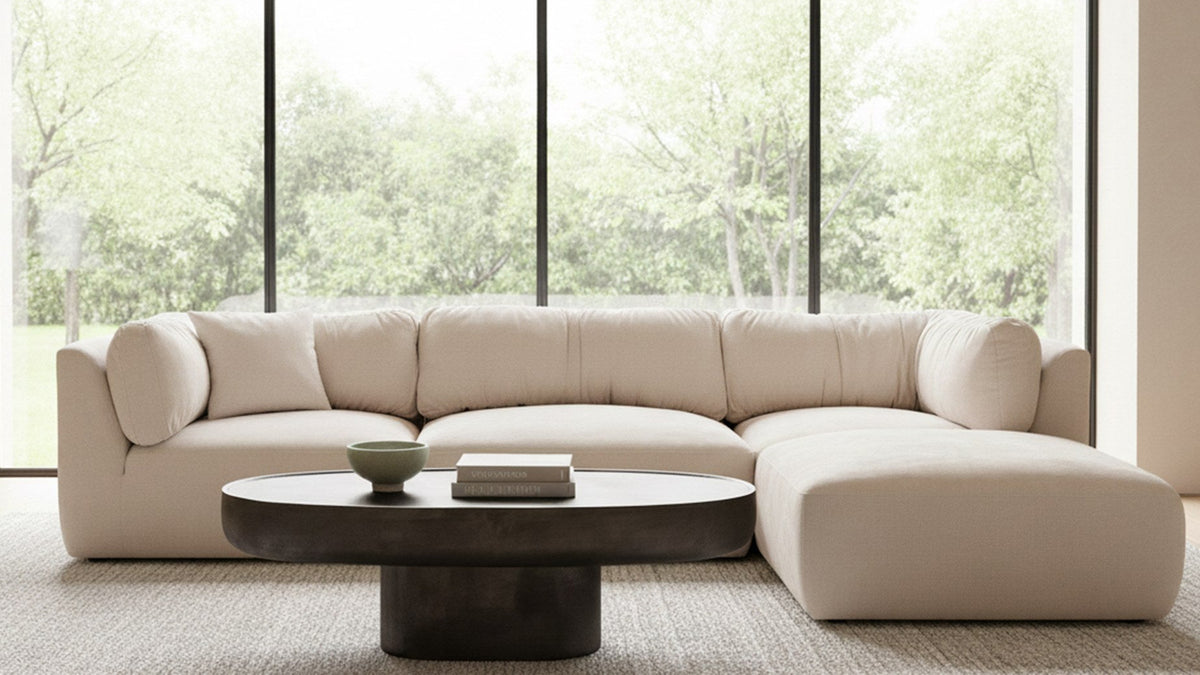 Moe's Home Collection Walter coffee table in deep bronze cast aluminum, its oval top carried on a single pedestal base, standing on a pale wool rug in front of a cream curved sectional sofa, with floor-to-ceiling windows onto a green garden behind, styled with a pale green bowl and two stacked art books