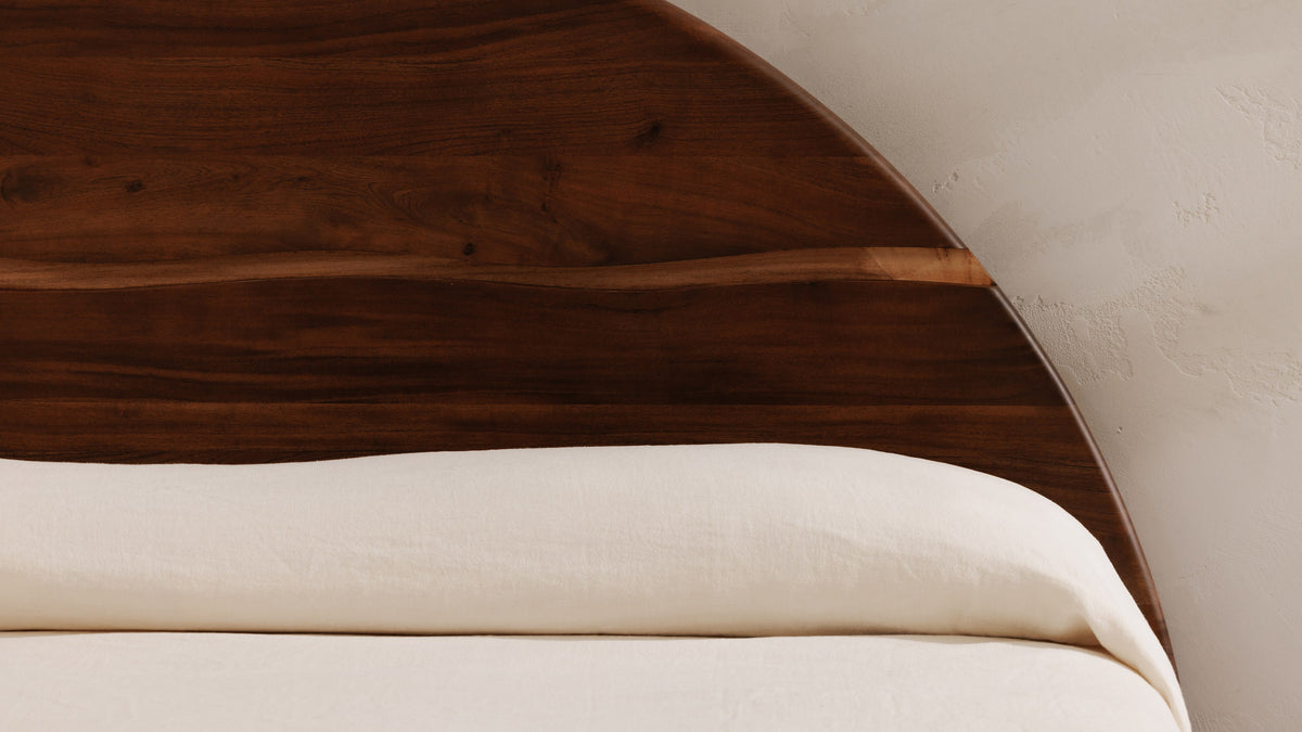 Close view of the Watson bed's arched headboard, showing the live edge running across the walnut-finished acacia above cream bedding
