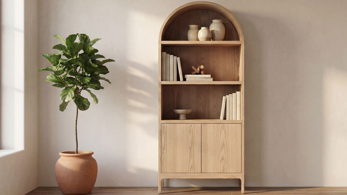 Moe's Home Collection Wilde display cabinet in natural oak, the arched top holding ceramics and books above two closed doors, beside a potted fig