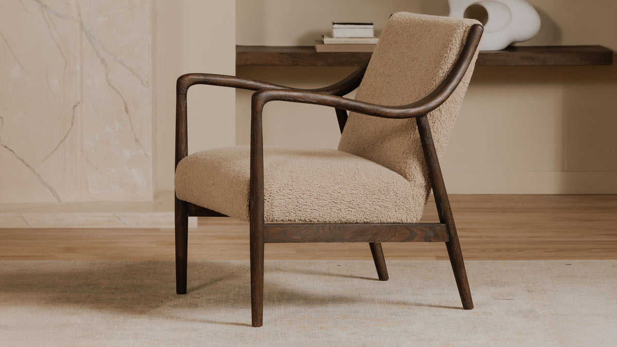 A Moe's Home Collection Yates accent chair in beige faux shearling on a deep brown solid oak frame, beside a walnut console holding stacked books against a marble wall